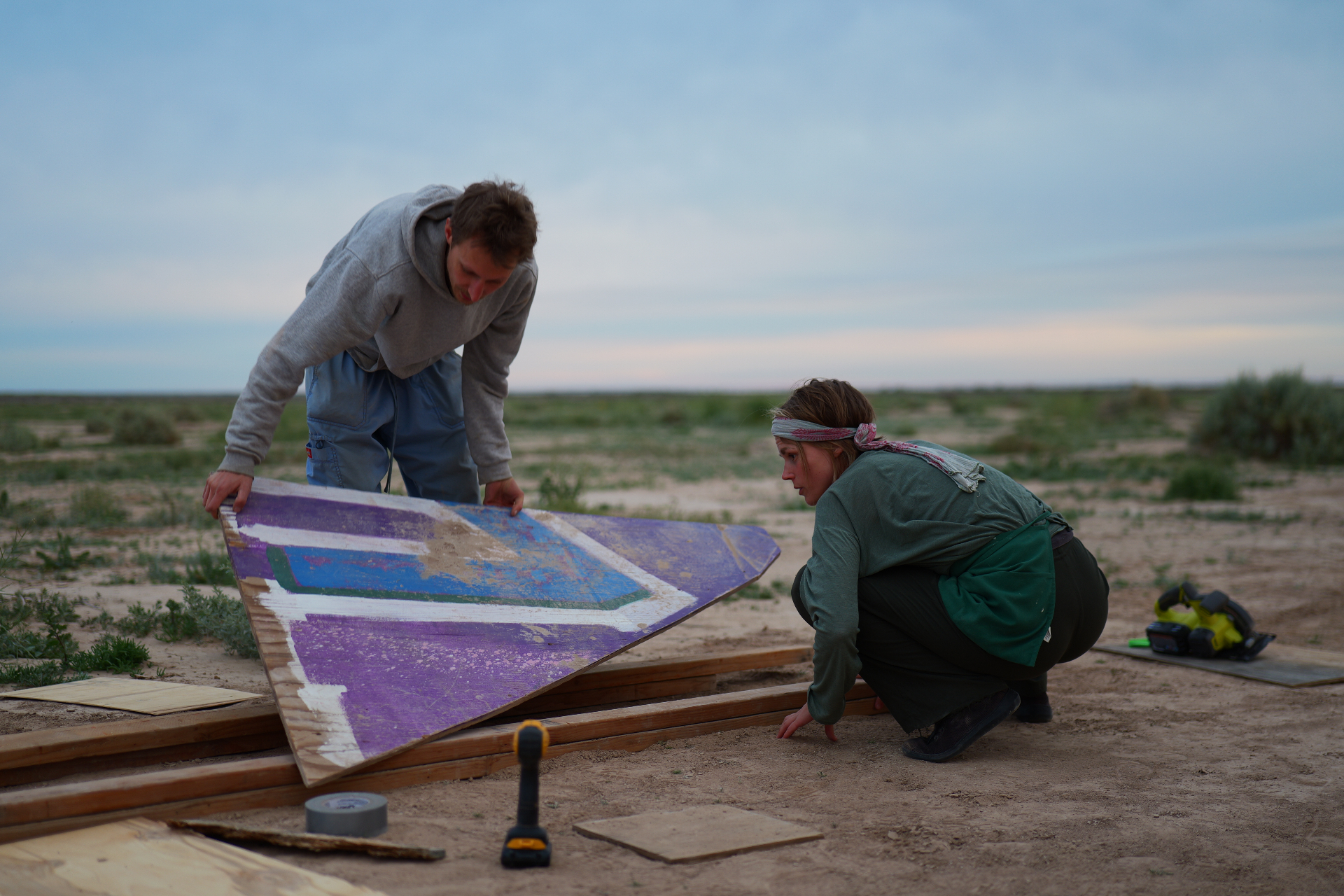 Two campmates inspecting a painted plywood panel at dusk